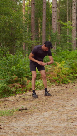 Video-Vertical-De-Un-Hombre-Haciendo-Ejercicio-Al-Aire-Libre-Corriendo-Por-Una-Pista-En-El-Bosque,-Deteniéndose-Para-Tomar-Aire-Y-Verificando-La-Aplicación-De-Actividad-En-Un-Reloj-Inteligente,-Filmado-En-Tiempo-Real-1