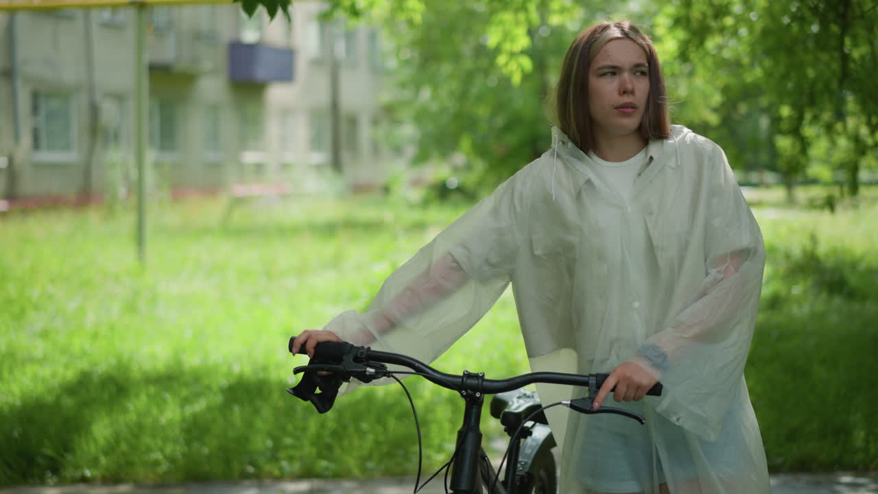 반투명한 레인코트를 입은 금발의 여성은 자전거를 타고 포장된 길을 산책하면서 피곤해 보이는데, 오른손에 검은 물건을 들고, 배경에는 울창한 초록색, 나무와 건물이 있습니다.