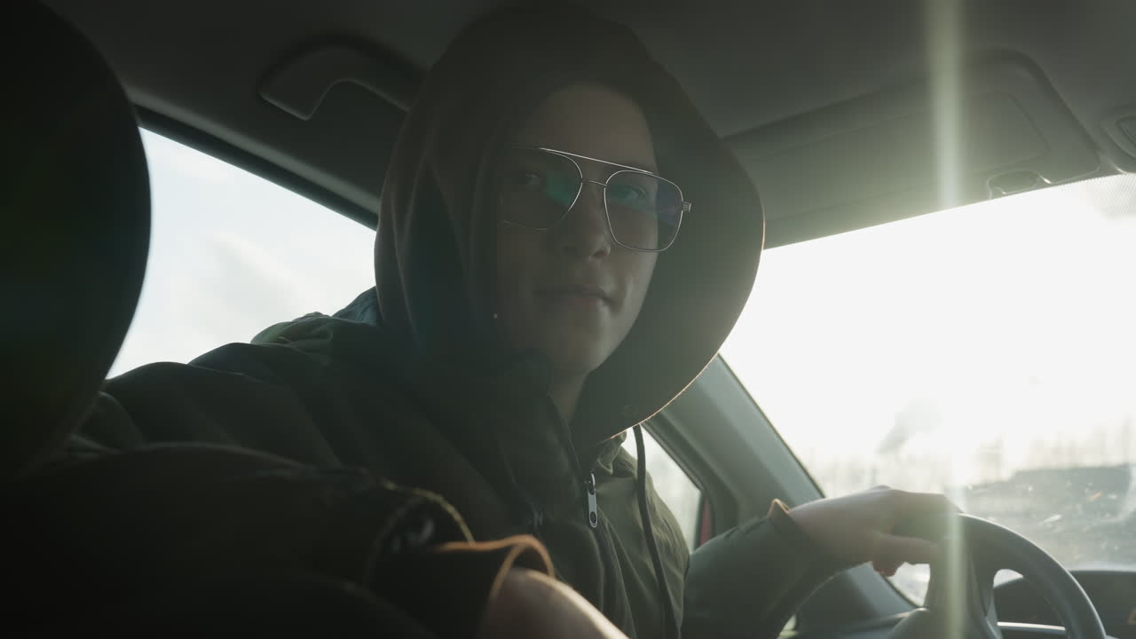 student wearing hoodie and tinted glasses gazing confidently over shoulder while driving car interior illuminated by sun flare through windshield reflecting winter parking lot beyond