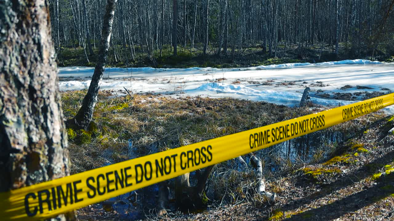 Yellow crime scene tape with the words do not cross stretches across a desolate forest landscape during early spring, with a frozen, melting body of water visible in the background