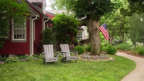 static shot of 2 adirondack chairs in the front yard of a red house with a betsy ross american flag in the background