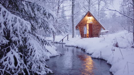 romantic honeymooners in towels exit wooden sauna near stream into snowy woods