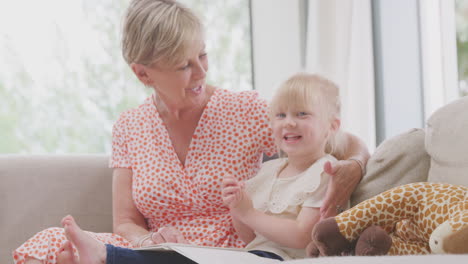 grandmother sitting on sofa with granddaughter at home reading book together