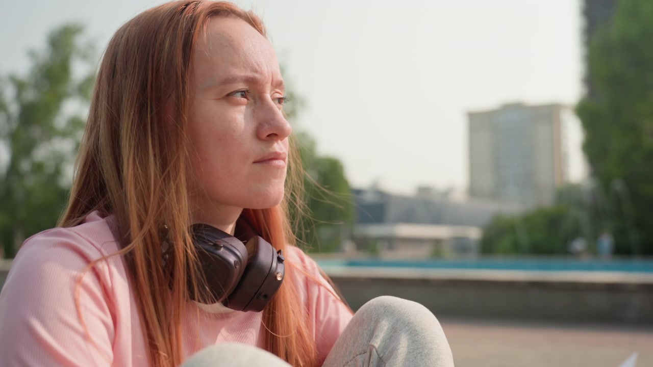 Una joven con cabello largo se sienta al aire libre en un día soleado, llevando auriculares alrededor del cuello y sosteniendo un papel. La mujer se encuentra en un parque urbano con árboles, edificios y una suave luz diurna creando un ambiente relajado.