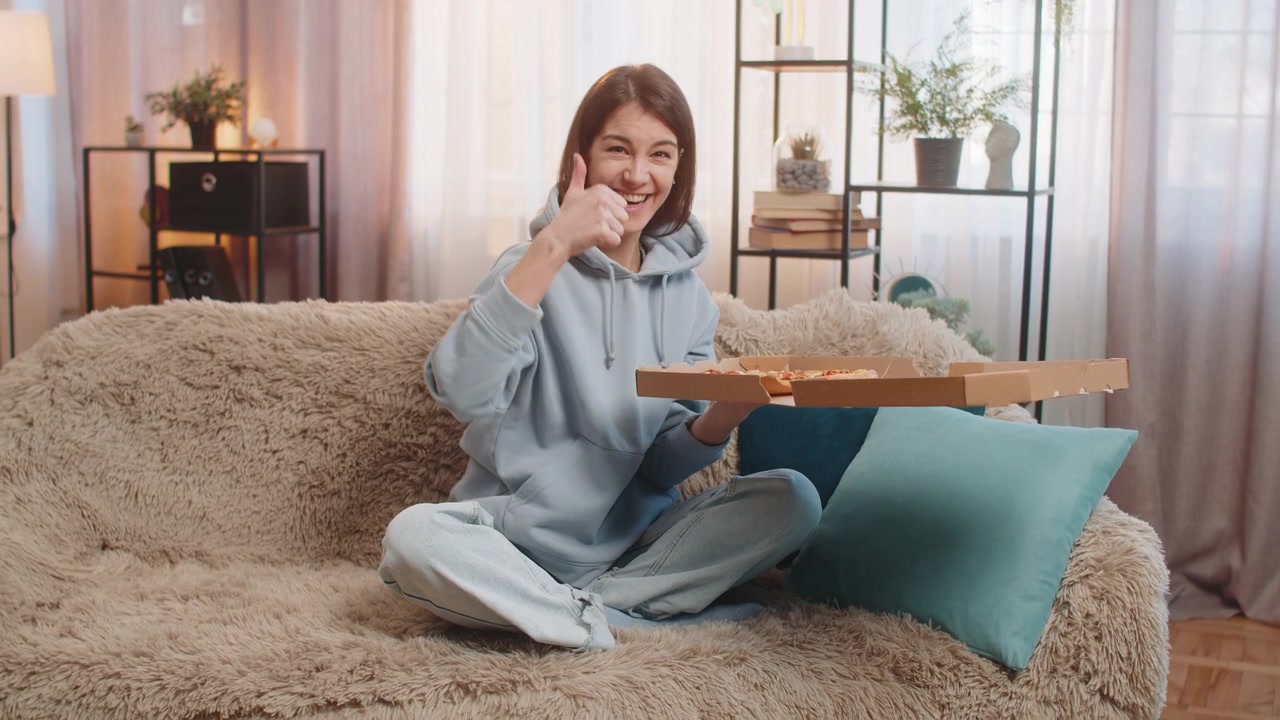 행복한 젊은 백인 여성이 배달 상자를 받아 포장을 풀고 집에서 맛있는 피자를 즐기고 있습니다.