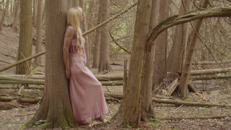 joven mujer de ébano se apoya contra el tronco de un árbol de goma de azúcar, en el bosque, ella está relajada, disfrutando de las vistas que la rodean. ella tiene el cabello rubio ondulado largo, con un vestido rosa, 60 fps de cámara lenta.
