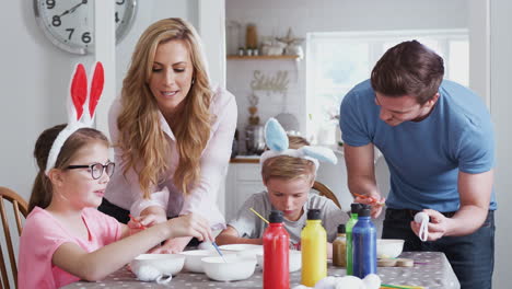 padres con niños que llevan orejas de conejo sentados en la mesa decorando huevos para la pascua en casa