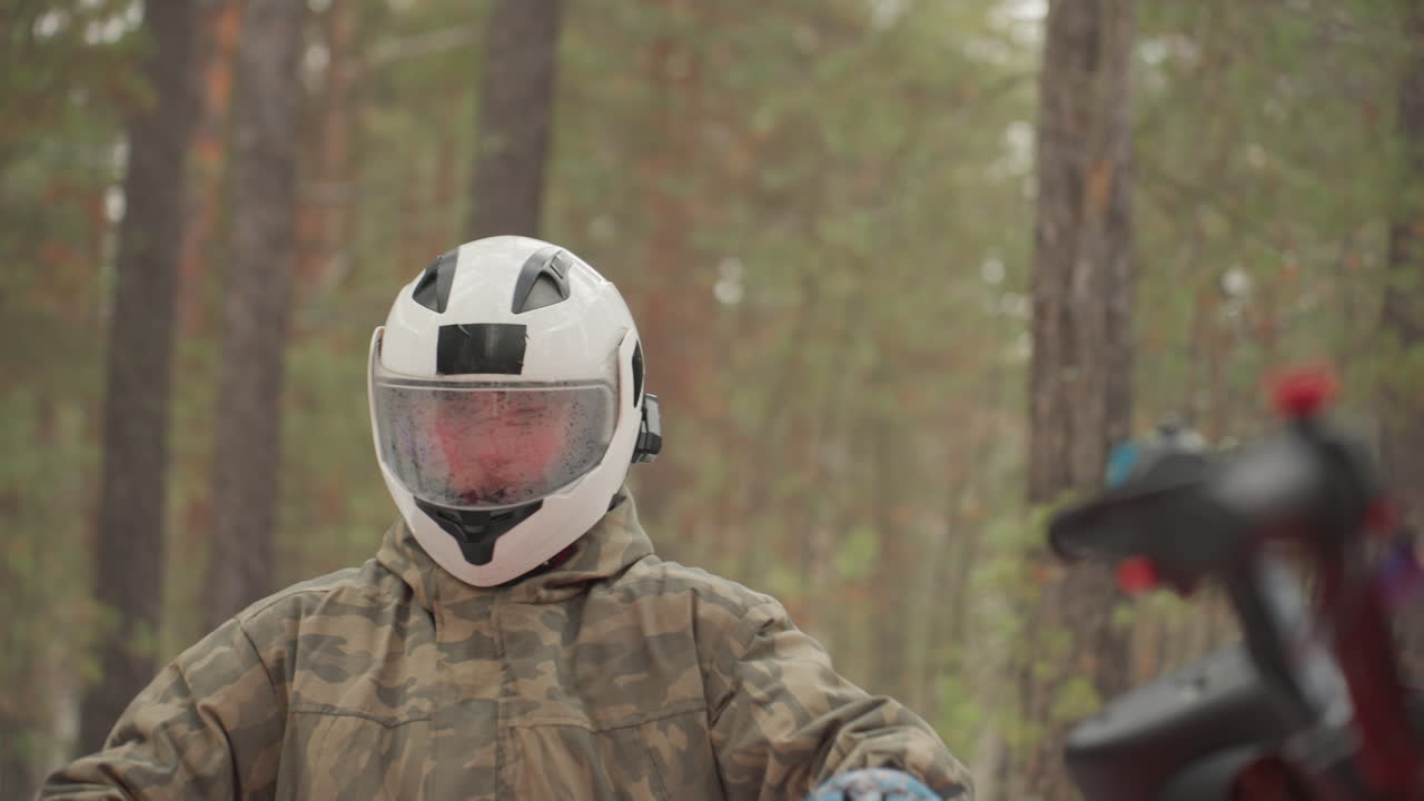 Un pilota si trova nella foresta, indossando una giacca mimetica e un casco bianco. Si sistema i guanti, sale sull'ATV rosso e avvia il motore. C'è una leggera nevicata, con pini sullo sfondo. Una preparazione concentrata per la guida fuoristrada.