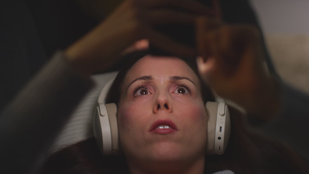 primer plano de una mujer con auriculares inalámbricos acostada en el sofá en casa por la noche transmitiendo o viendo contenido en línea en el teléfono móvil