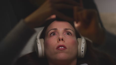 close up of woman wearing wireless headphones lying on sofa at home at night streaming or looking at online content on mobile phone