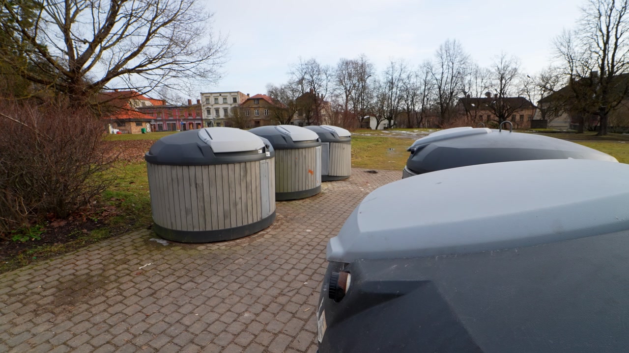 Row of underground municipal waste dumpsters in an urban park on a paved walkway, with leafless trees and overcast sky—public sanitation and neighborhood trash collection.