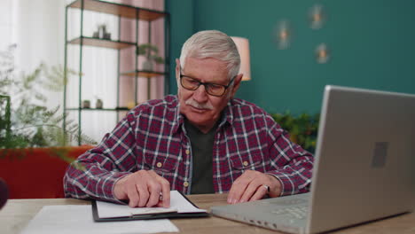 senior man working on laptop computer, making notes on sheet of paper, upset by poor bad results