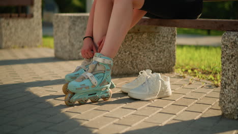 partial view of individual adjusting roller skate straps on right leg while seated , with white sneakers placed behind on pavement, greenery in the background, warm sunlight casting gentle shadows