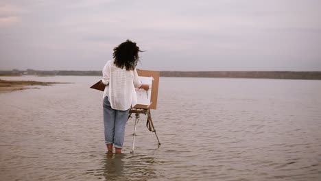 The-female-artist-in-blue-jeans-and-white-shirt-standing-with-easel-in-the-water-till-ankles-and-working-on-her-future-picture.-Beautiful-surrounding-landscape:-lake-and-clear-white-sky.-Backside-view