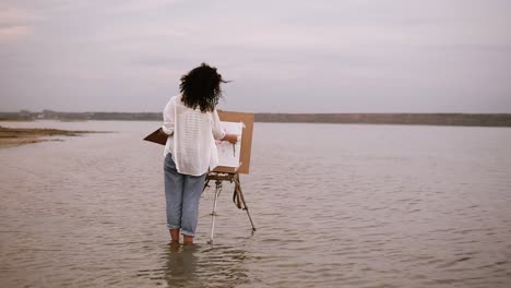 the female artist in blue jeans and white shirt standing with easel in the water till ankles and working on her future picture. beautiful surrounding landscape: lake and clear white sky. backside view