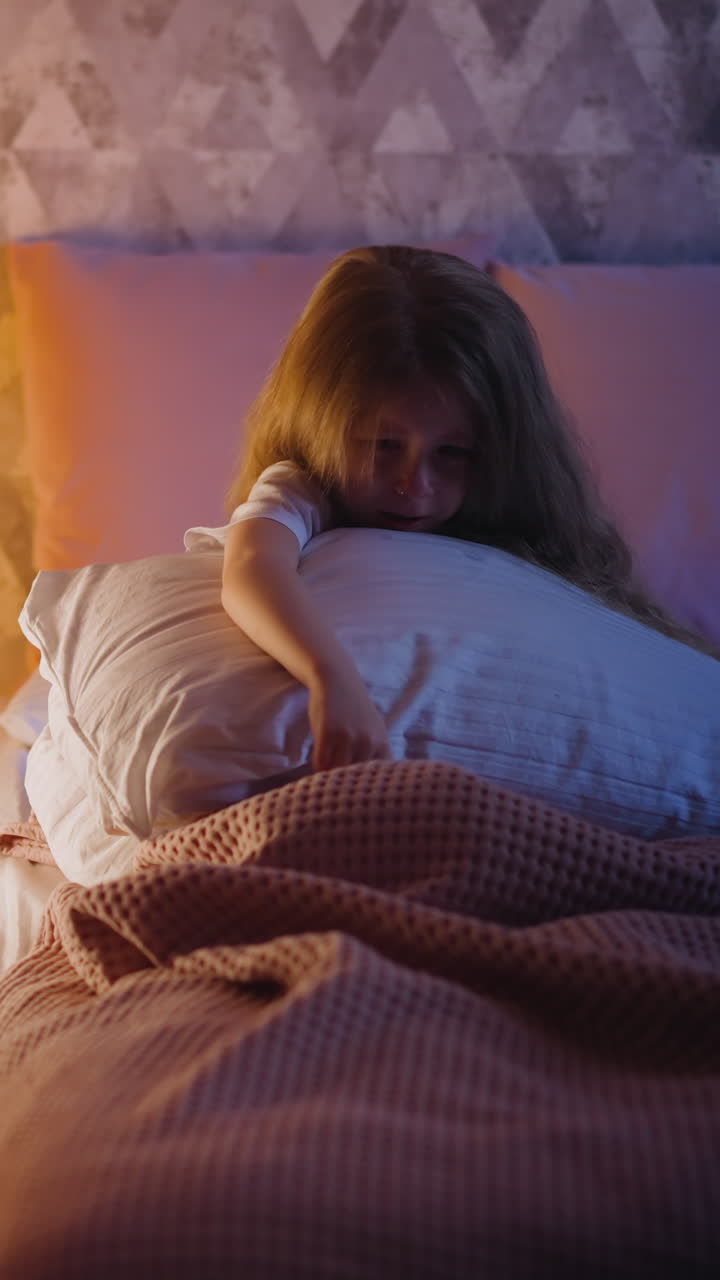 Upset blonde little girl hugs large pillow crying on bed near children wigwam with glowing lamp inside near grey wall in bedroom at night slow motion