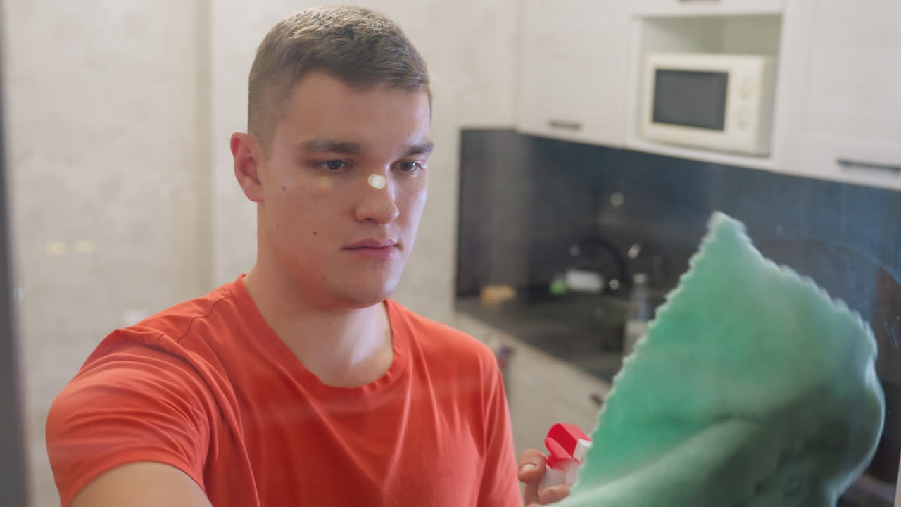 Man in orange shirt holding spray bottle in front of mirror, applying cleaning solution with concentration, removing water streaks and marks, focusing on household hygiene