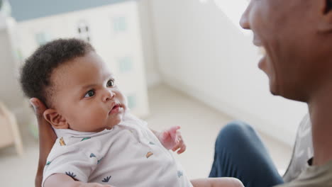 Over-The-Shoulder-View-Of-Father-Cuddling-And-Playing-With-Baby-Son-In-Nursery-At-Home