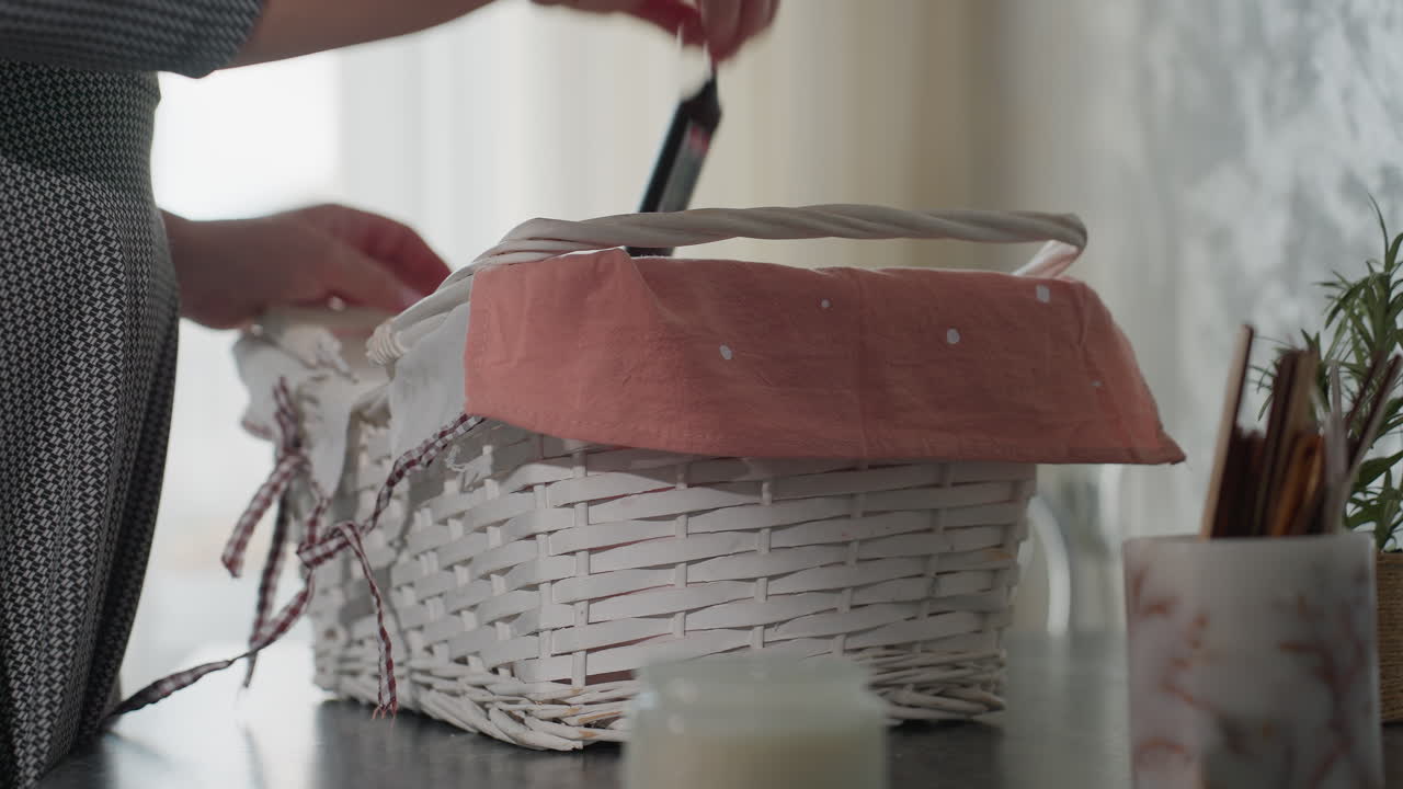 Close up partial view of woman in checkered outfit returning from shopping placing white basket with pink cloth liner on kitchen table and picking item from inside under soft daylight interior