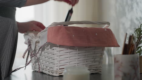 close up partial view of woman in checkered outfit returning from shopping placing white basket with pink cloth liner on kitchen table and picking item from inside under soft daylight interior