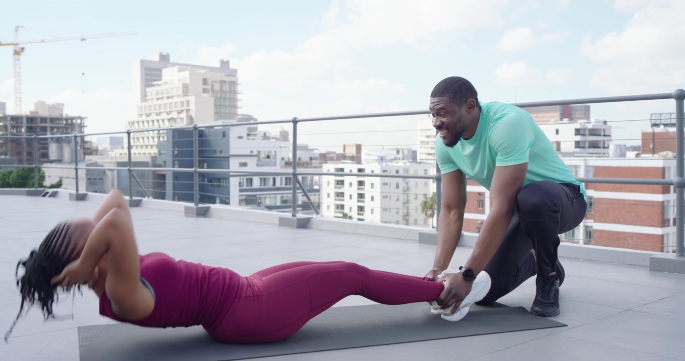 Coach afro-américain tenant des chaussures, donnant des instructions à une cliente en marron faisant des abdominaux sur un tapis pour le gainage.
