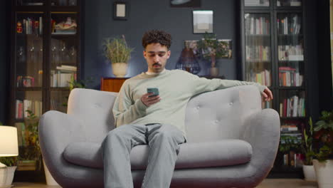 Young-Man-Using-Smartphone-While-Sitting-On-A-Couch-At-Home