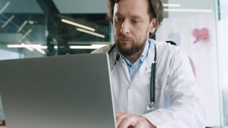 close-up view of senior caucasian male doctor sitting in hospital office working and typing on laptop at workplace