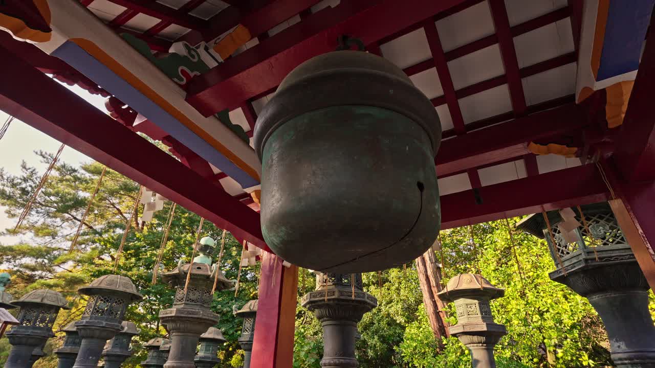 A weathered bronze bell hangs beneath a red, traditional Japanese structure, surrounded by ancient stone lanterns. This close-up highlights the historical and spiritual essence of the shrine.