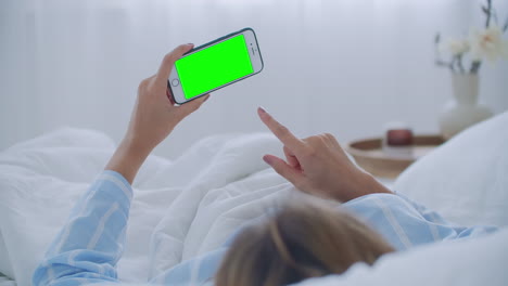 침대 어깨에  ⁇ 드폰을 든 젊은 여성. 기술 중독. 녹색 화면 전화. 침실에서 배를 만지고 스마트폰을 사용하는 젊은 여성