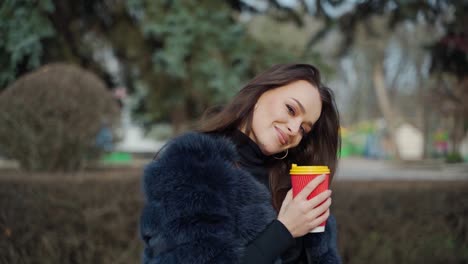 portrait of a smiling woman. attractive brunette girl in fur coat stands with plastic cup of coffee in the park and looking on camera. slow motion.