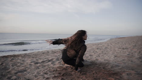 Mujer-Rubia-Con-Un-Vestido-De-Tela-Negra-Sexy-Bailando-Sola-En-La-Playa-De-Arena-Al-Atardecer-Con-Las-Olas-Del-Océano-En-El-Fondo