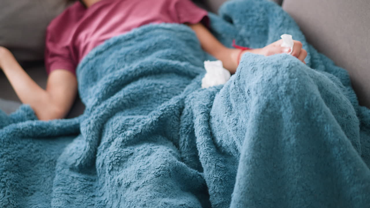 Close-up of weak woman adjusting herself under soft blue blanket, surrounded by tissues, showing signs of illness and fatigue, indicating cold or flu symptoms with tissue on her