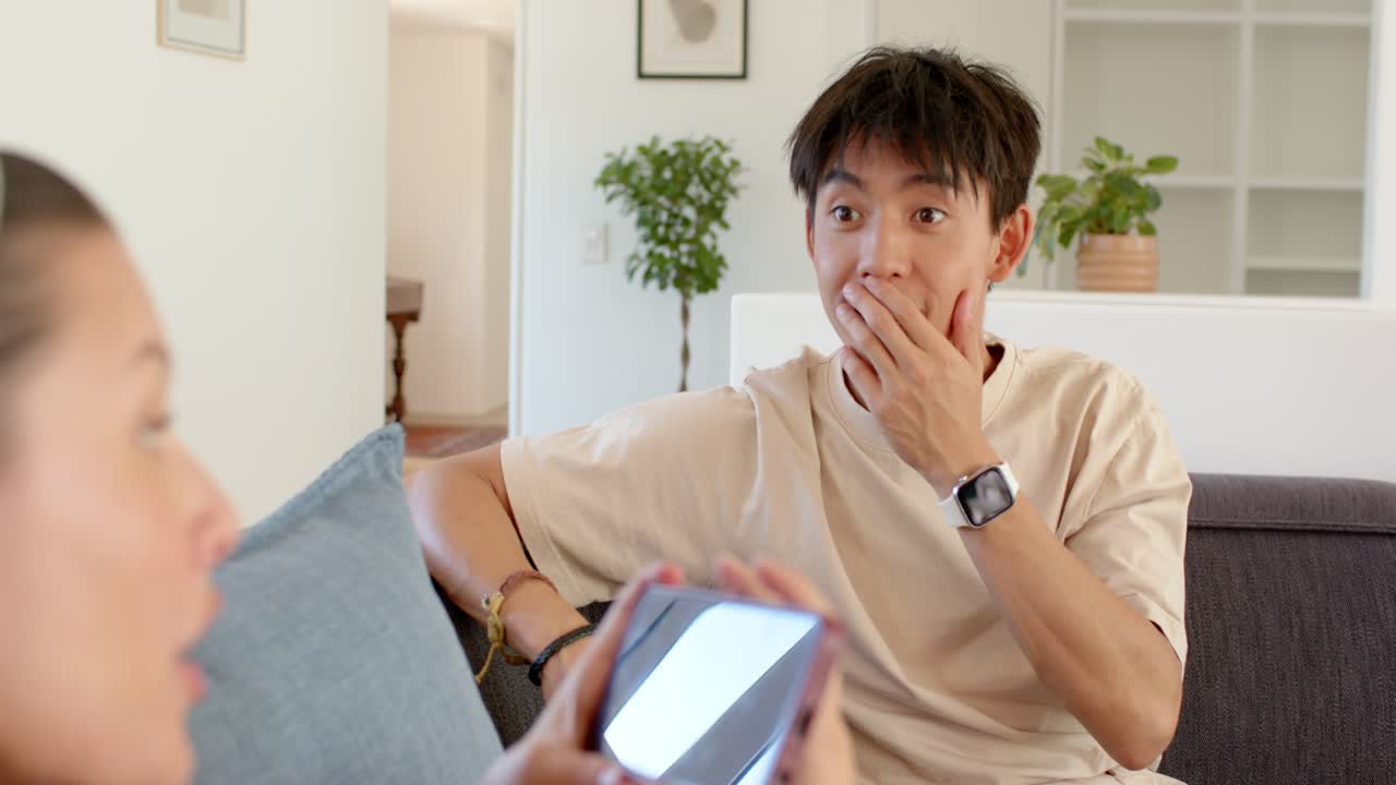 ソファに座るカップル。女性が赤いケースのスマートフォンを見せ、男性はそれに反応し、タブレットとスマートフォンを見ようと身を乗り出している。