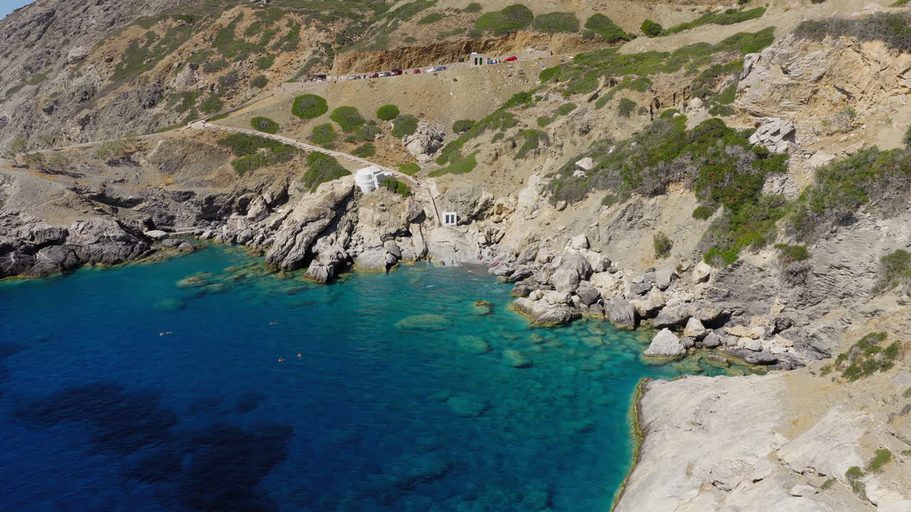 Aerial panoramic approach to Agia Anna beach, Amorgos island. People swimming in blue waters below the hillside church