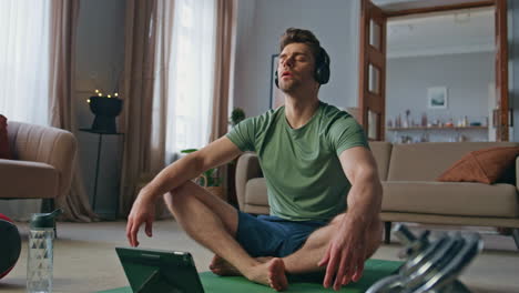 serene athlete meditating headphones at apartment. calm fit man sitting in lotus
