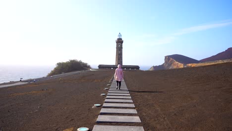 woman in pink jacket walks towards lighthouse of ponta dos capelinhos surrounded by volcanic ashes, rear shot