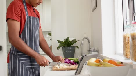 hombre biracial en delantal cortando verduras y usando tableta en la cocina, cámara lenta