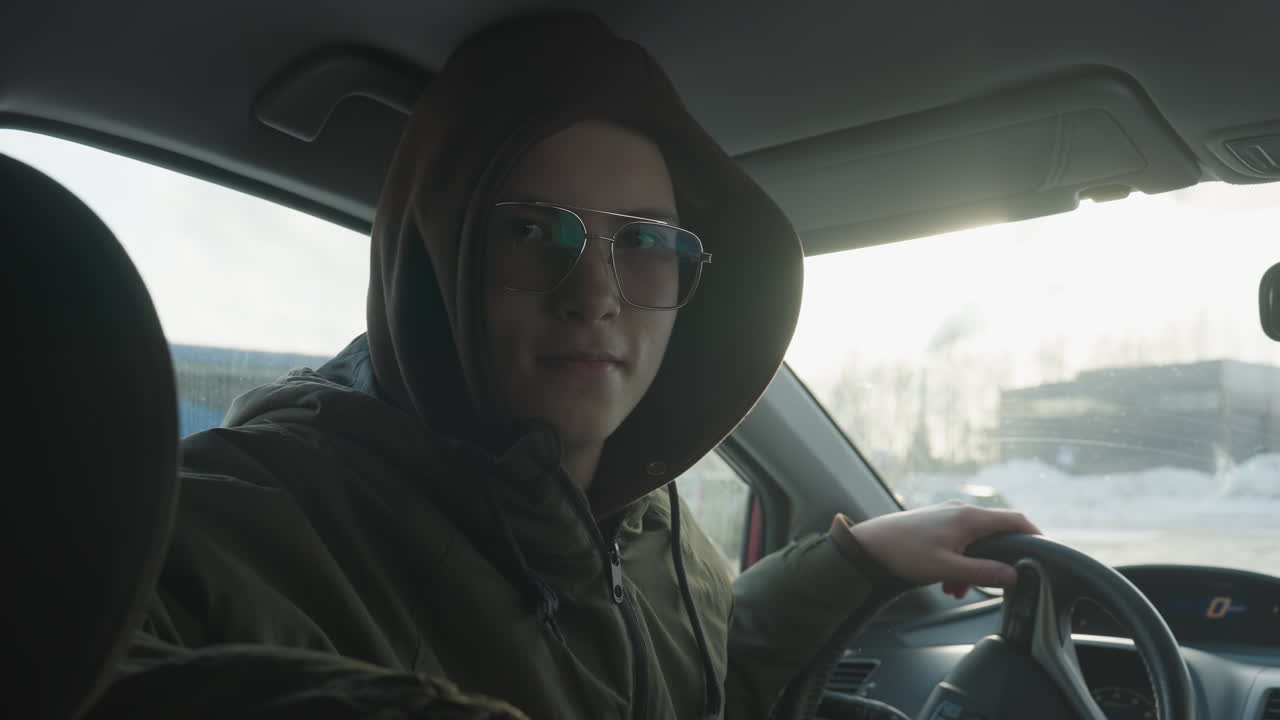 young boy in hoodie gripping steering wheel with confident focus tinted glasses reflecting winter landscape beyond windshield moody cabin lighting casting subtle shadows on face inside parked car