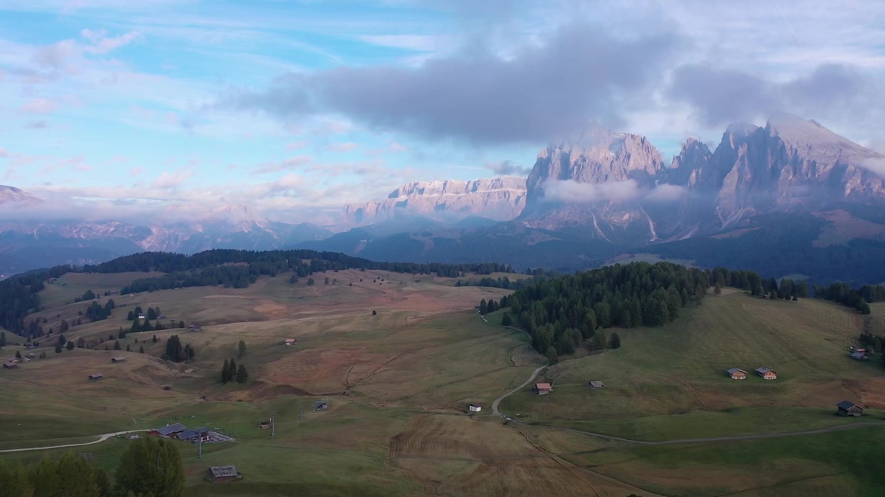 Betagende droneoptagelse fra høj vinkel af de bølgende grønne græsgange og alpehytter ved Alpe di Siusi i de italienske Dolomitter. Med de majestætiske Sassolungo-bjergtoppe under overskyet sommerhimmel.