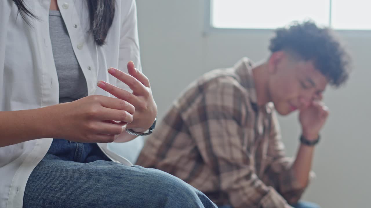 Esposa tirando a aliança de casamento com homem frustrado ao fundo.