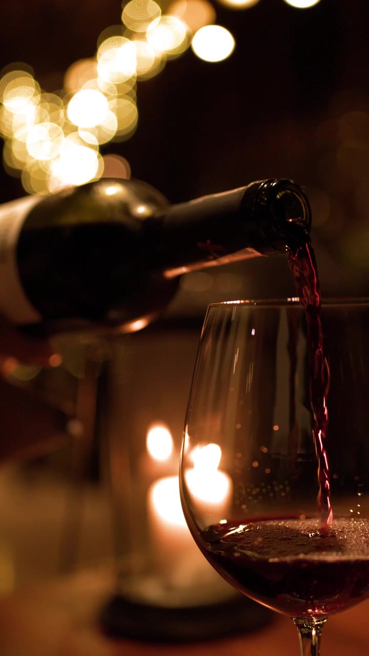 Vino rosso che si versa in un bicchiere sotto la calda luce di una candela con una morbida illuminazione bokeh sullo sfondo.