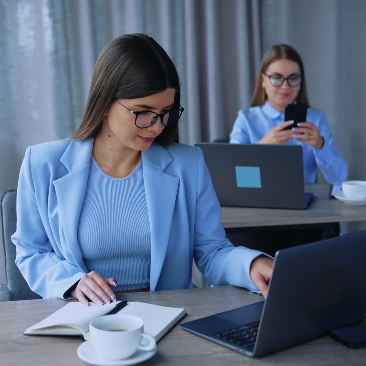 Young female ladies sitting at desks in front of laptops. Employees using gadgets in modern office. Blurred backdrop