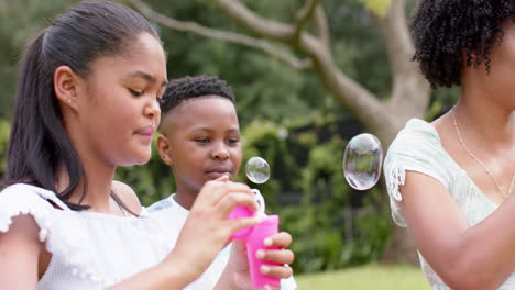 Glückliche-Afroamerikanische-Mutter,-Tochter-Und-Sohn-Pusten-Seifenblasen-Im-Garten,-Zeitlupe