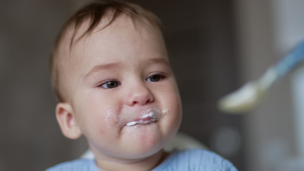 Adorable grey-eyed baby boy in blue shirt being fed from spoon. Lovely child eating dairy portrait close up.