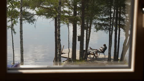 man relaxes on a lakeside dock reading a book under pine trees, seen through a window in summer