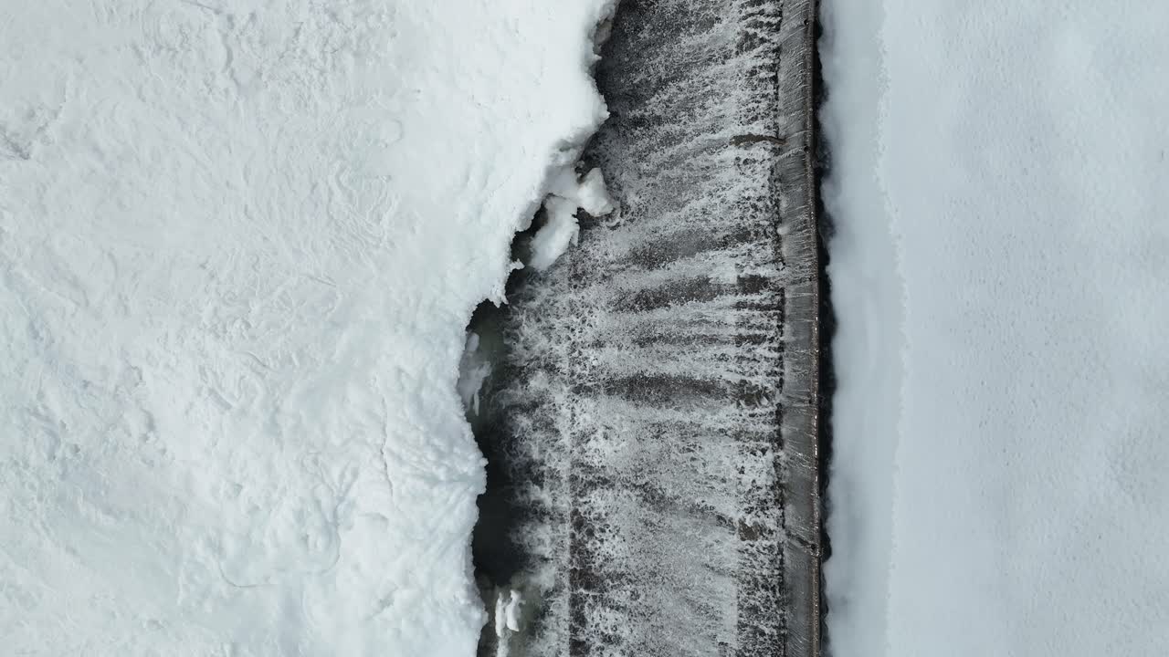 A drone captures a top-down view of a dam during the winter, moving from top to bottom, showcasing water flowing over the retaining boards and the ice structures formed as a result.