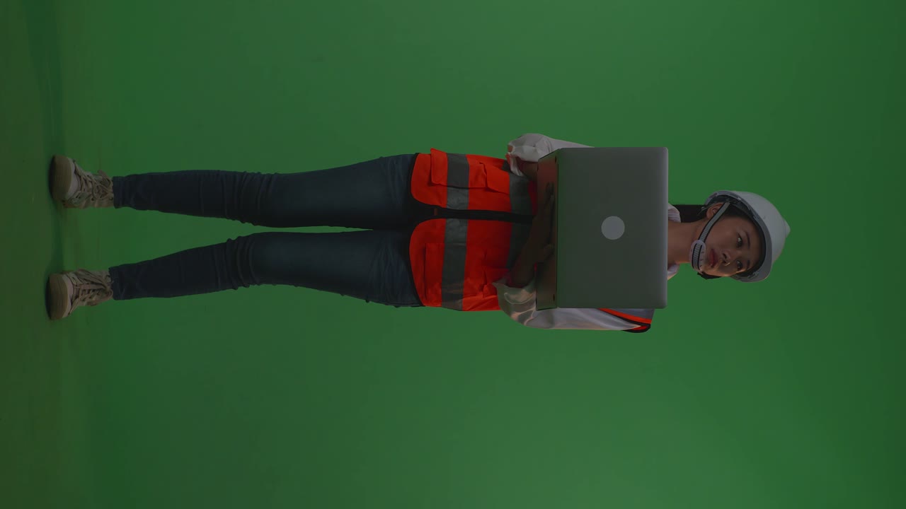 cuerpo lleno de ingeniera asiática con casco de seguridad trabajando en una computadora portátil y mirando a su alrededor mientras está de pie en el estudio de fondo de pantalla verde, durante la puesta o el amanecer
