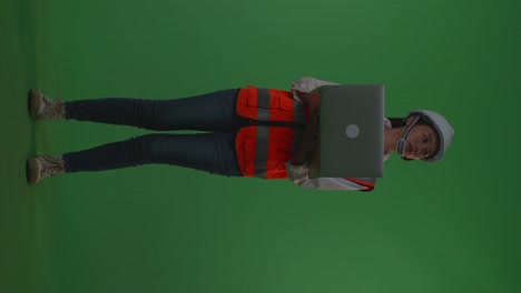 full body of asian female engineer with safety helmet working on a laptop and looking around while standing in the green screen background studio, during sunset or sunrise time