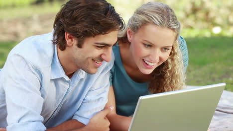 una pareja sonriente yace en el parque con una computadora portátil mientras miran hacia la cámara