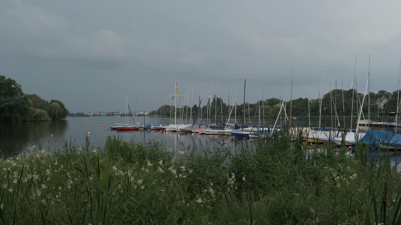 Streak of forked lightning over the city of Hamburg shrouded in grey cloud as seen from the Outer Alster lake with moored boats at a jetty over greenery in the foreground